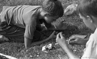 Photographie en noir et blanc montrant deux garçons accroupis dans l’herbe, l’un essaie de s’allumer une cigarette.