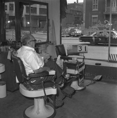 Un homme est assis à l’intérieur du salon de coiffure Barbier A. Plouffe et regarde à l’extérieur par la grande fenêtre.