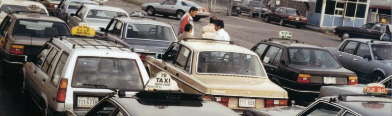 Photo couleur montrant plusieurs taxis de différentes compagnies dans un stationnement. Quelques hommes sont regroupés près d’une des voitures. 