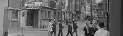 Scène de rue en noir et blanc dans le Quartier chinois de Montréal. Des hommes et des femmes traversent la rue ou marchent sur le trottoir. On voit des façades avec des enseignes commerciales en chinois.