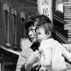 Scène de rue en noir et blanc. Une femme tenant un bébé dans ses bras marche dans le Quartier chinois. On voit des façades avec des enseignes commerciales en chinois.