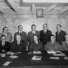 Photographie en noir et blanc d’un groupe d’hommes se tenant derrière une table de travail. Ils portent tous un complet-cravate.