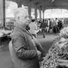 Photo en noir et blanc d’un homme âgé se tient devant un kiosque de légumes dans un marché. 