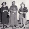 Un homme d’Église est entouré de deux femmes, une habillée d’un costume et l’autre avec fourrure. Les trois ont un sourire, mais la femme de droite sourit plus amplement en regardant les deux autres.