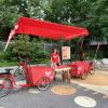 Devant un parc, une femme se tient debout derrière deux vélos dotés de grands coffres et d’une tente de couleur rouge.