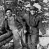 Photographie de deux hommes appuyés sur des billots de bois, dans la forêt en hiver.