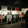 Salle d’exposition montrant deux murs avec des photos en noir et blanc, des panneaux de texte, un écran et deux personnes, debout, qui la visitent. 