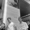Deux militants anti-guerre du Vietnam portent des chandails avec des messages, l’un écrit « Stop the bombs » et l’autre « Peace in Vietnam ». Un homme à leur gauche leur montre un document. 