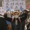 Une foule marche dans une rue du centre-ville. Plusieurs personnes tiennent des pancartes ou des bannières en chinois ou en anglais.