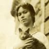 Jeune femme, en robe, tenant un chat dans ses bras, sur un balcon d’appartement.