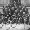 Environ 35 hommes et garçons en rangée posent devant un édifice, avec des vélos en avant-plan.