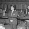 Salle de tribunal remplie d’hommes. À gauche, un homme en complet cravate se tient debout derrière une balustrade. Il a les mains croisées devant lui. 