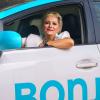 Photo couleur format portrait d’une chauffeuse de taxi souriant au photographe par la fenêtre ouverte de la portière. 