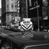 Photo en noir et blanc montrant la rue Sainte-Catherine à la hauteur des rues Peel et Drummond en 1969. On y voit des passants sur le trottoir, les enseignes des magasins, et une voiture de taxi dans le coin inférieur droit de la photo avec un lanternon V