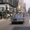 Photo couleur montrant la rue Sainte-Catherine, au centre-ville de Montréal, avec des voitures et des autobus qui circulent dans la rue, une foule au coin de la rue Peel, et les édifices de chaque côté.  