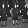 Photographie en noir et blanc d’un groupe de femmes en uniforme dans une salle lambrissée. 