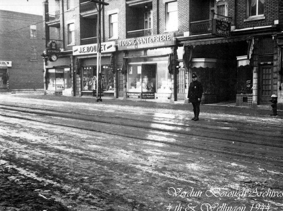 Photographie noir et blanc de la rue Wellington au premier plan et, en arrière-plan, du commerce Tousignant & Frères. Un policier se tient debout dans la rue sur la droite. 