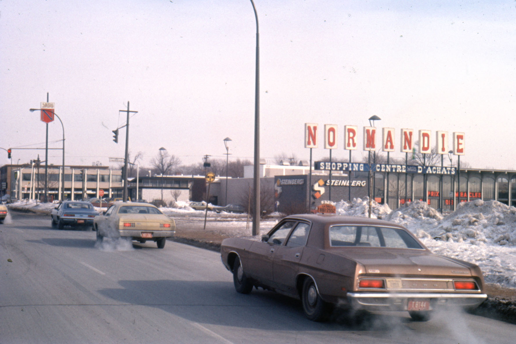 Photo couleur montrant un taxi et d'autres véhicules circulant dans une rue assez large. En arrière-plan, une enseigne annonce « NORMANDIE. SHOPPING CENTRE D’ACHATS » et on peut voir un supermarché Steinberg.