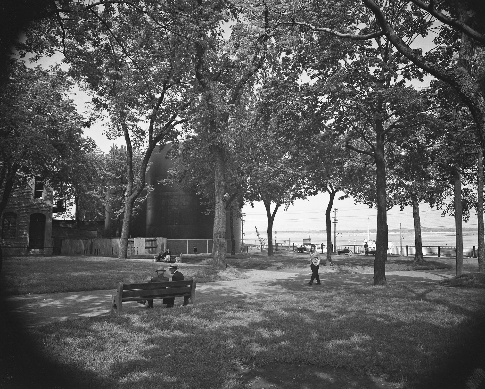 Photo en noir et blanc d’un parc avec de grands arbres sur le bord de l’eau. Deux hommes sont assis sur un banc et un autre homme marche. Des réservoirs à mélasse sont situés à l’arrière-planà gauche de la photo.