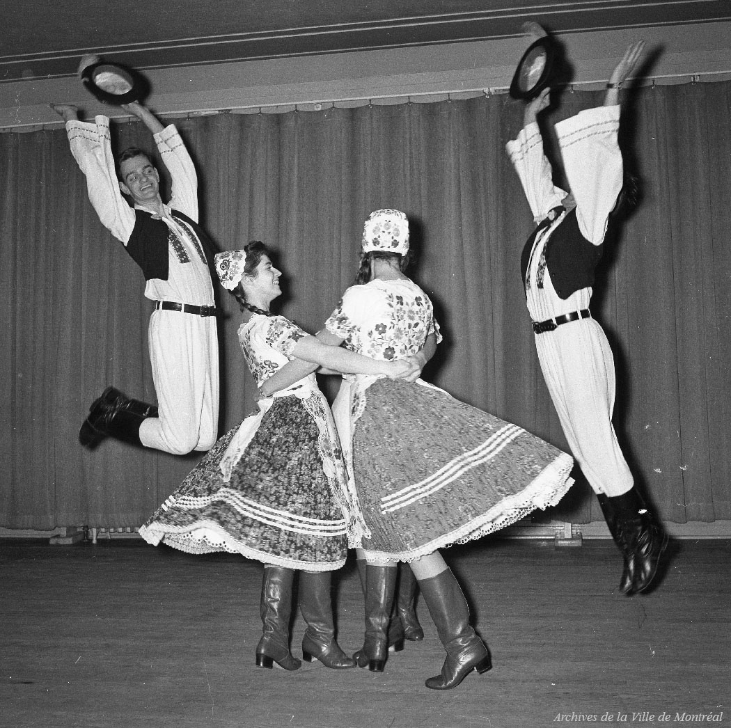 Deux danseurs sautent et sont de chaque côté de deux danseuses qui se tiennent par la taille. Ils sont tous en costume folklorique hongrois.