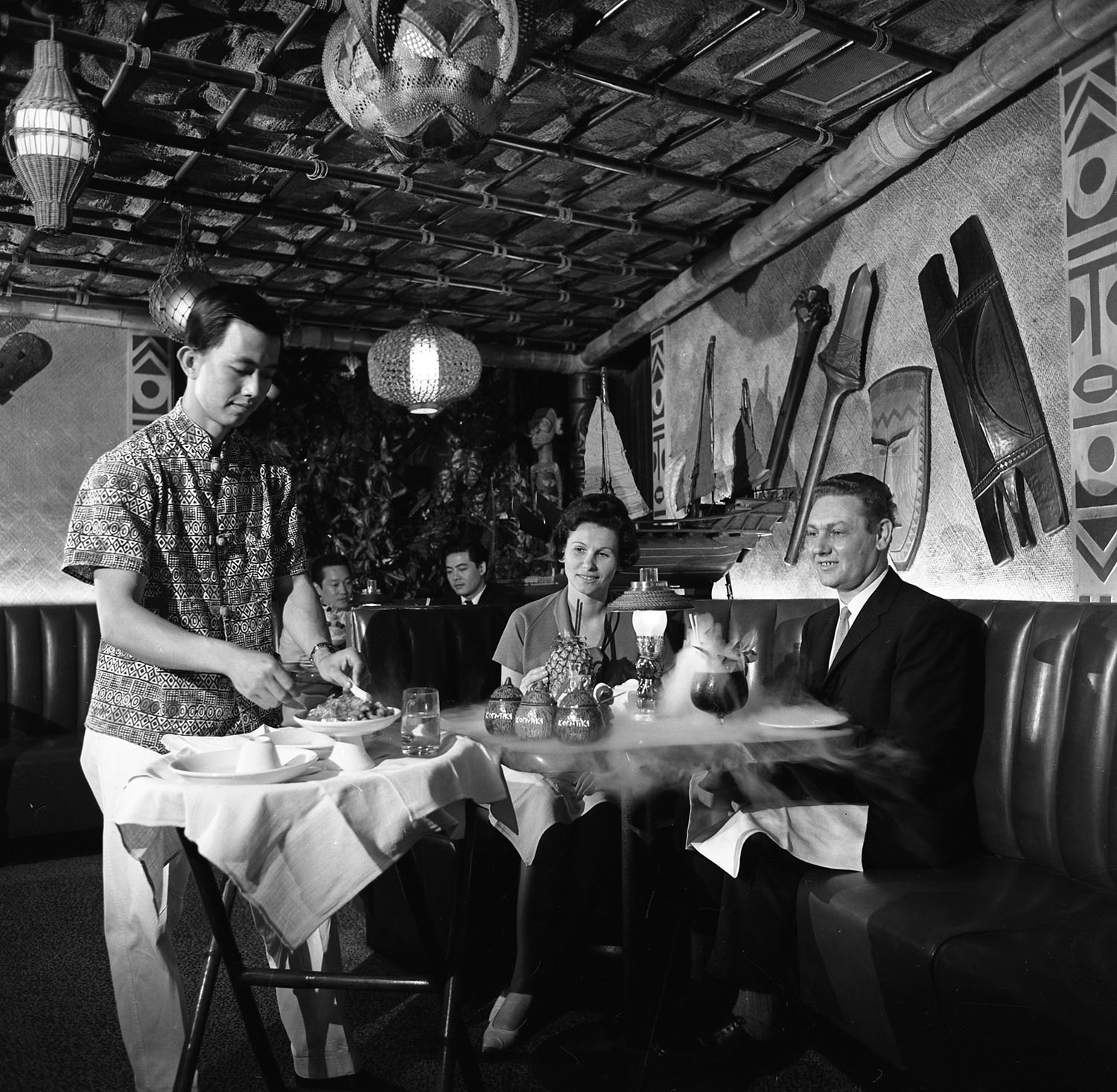 Photographie en noir et blanc de deux couples attablés. Le couple à l’avant est servi par un cuisinier et de la fumée s’échappe des verres sur la table. 