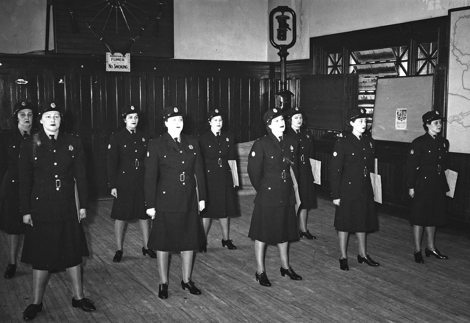 Photographie en noir et blanc d’un groupe de femmes en uniforme dans une salle lambrissée. 