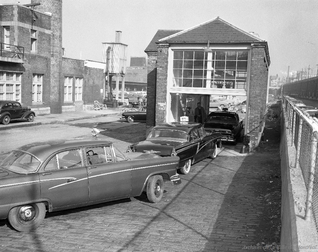 Photo en noir et blanc montrant deux voitures de taxi attendant à l’extérieur d’un garage, pendant qu’une troisième voiture est à l’intérieur pour une vérification. 