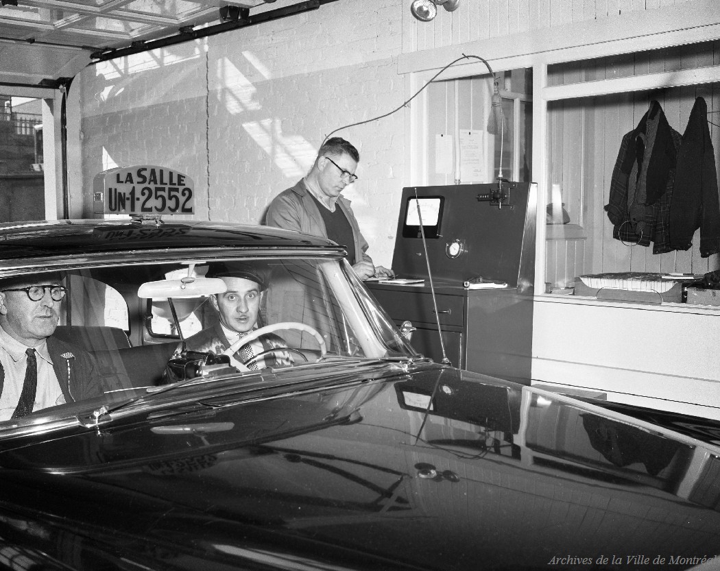 Photo en noir et blanc montrant en gros plan une voiture de taxi de la compagnie La Salle dans un garage. À l’intérieur de la voiture se trouvent le conducteur et un passager. Et à l’extérieur, un homme debout fait des vérifications à côté d’un appareil.