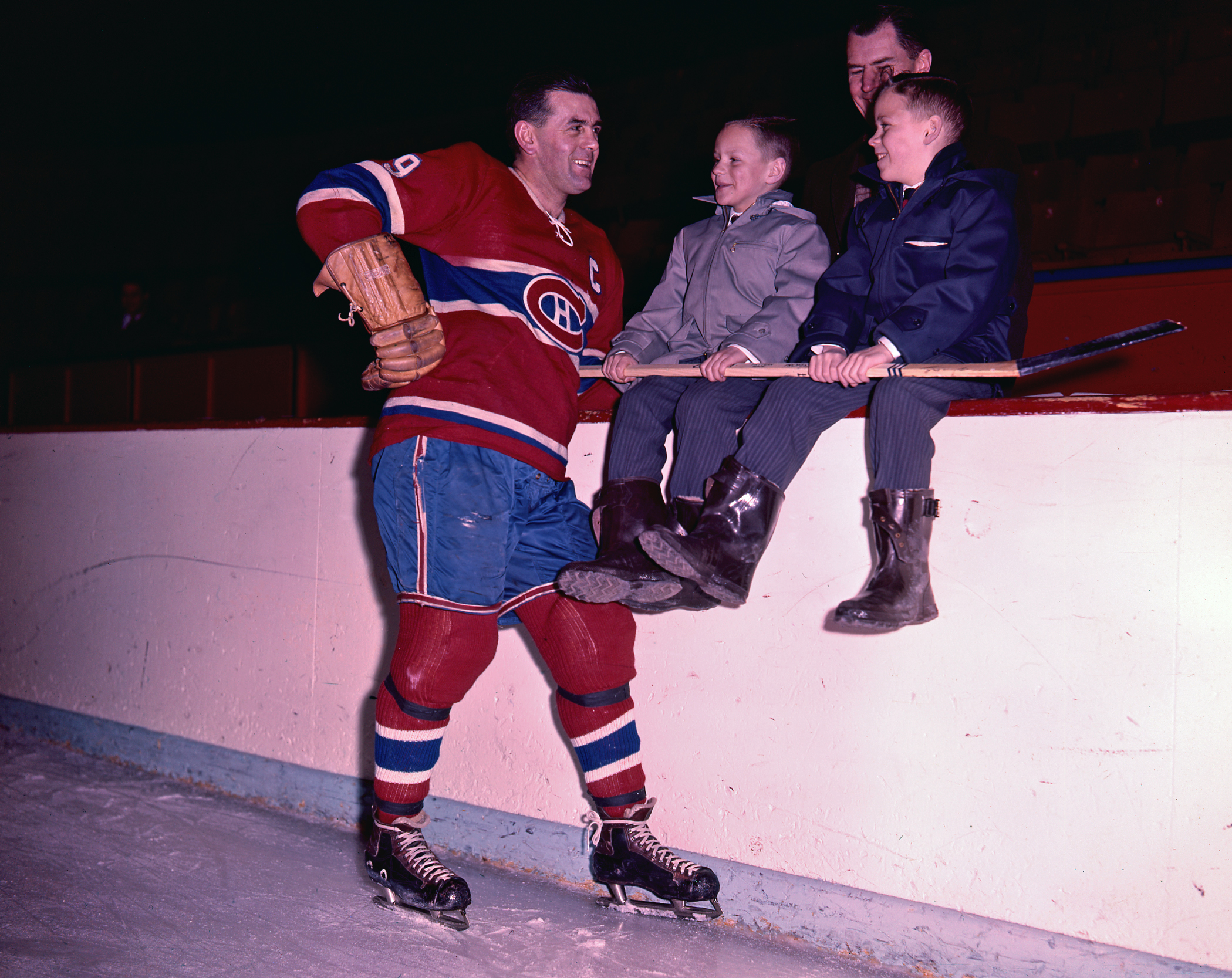 Photographie couleur de Maurice Richard affublé de son équipement du Canadien, souriant à deux jeunes garçons tenant un bâton de hockey.