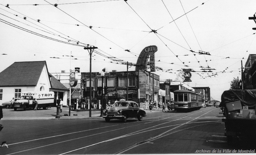 Photographie en noir et blanc. Une station-service Esso occupe le coin sud-est de l'intersection. En arrière-plan, on peut voir les enseignes d'un garage GMC et du Café Democ. Un tramway de la ligne 77 et un taxi remontent le boulevard.