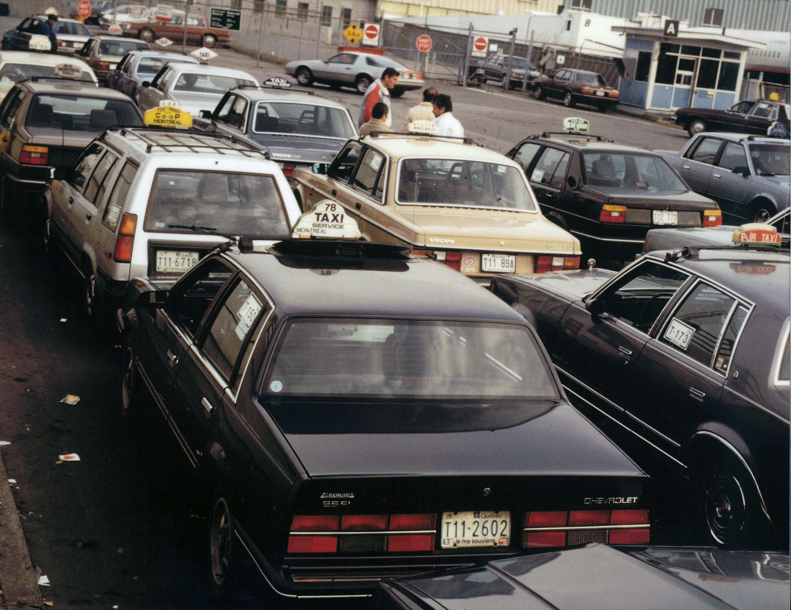 Photo couleur montrant plusieurs taxis de différentes compagnies dans un stationnement. Quelques hommes sont regroupés près d’une des voitures. 