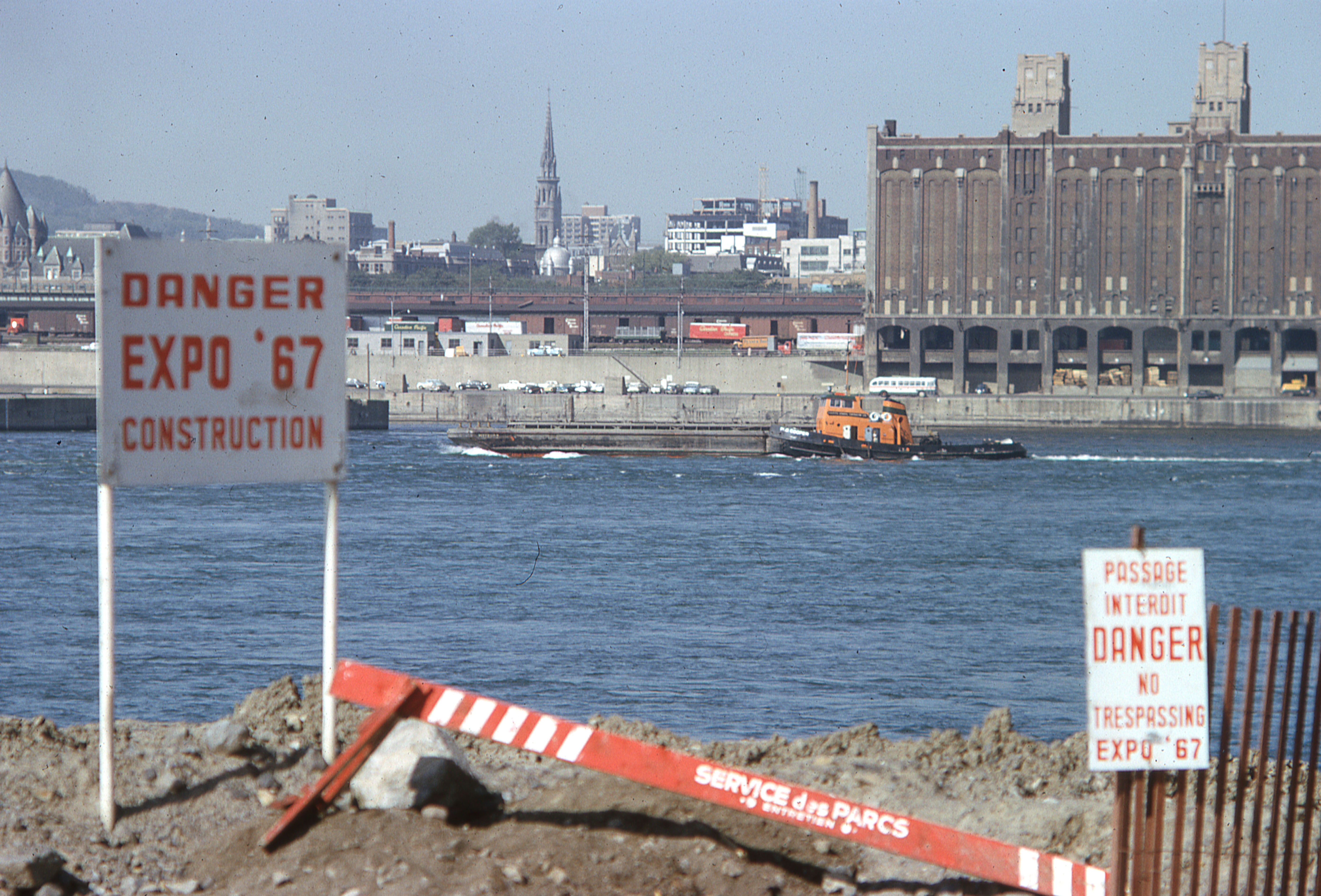 Vue montrant à l'avant-plan l'île Sainte-Hélène  avec un panneau disant "Danger Expo 67 Construction", avec Montréal à l'arrière-plan.