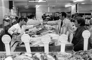 Photo en noir et blanc montrant l’intérieur d’une poissonnerie, avec quelques travailleurs et clients.