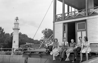 Six hommes et femmes sont assis sur la galerie devant un pavillon en bas. Un phare se trouve à gauche de la photo en noir et blanc.