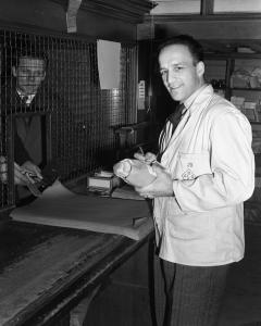 Photographie en noir et blanc d’un homme portant un sarrau avec le logo de la Commission des liqueurs et tenant une bouteille. Derrière la grille se tient un client.