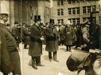 Photo d’un groupe d’hommes portant chapeau et costume sur le parvis d’un édifice (église?), des policiers sont présents.