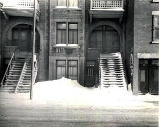 Photographie d’un bâtiment anonyme en pierre et en brique dont les entrées sont sous des arches. Il y a des escaliers qui partent du perron et donnent sur la rue.