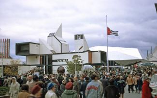Vue d’ensemble du pavillon cubain, avec le drapeau en berne, et de nombreux visiteurs font la file pour y entrer.