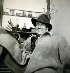 Un homme est assis. Il porte un manteau de drap et un chapeau de feutre, et a un verre à la main.