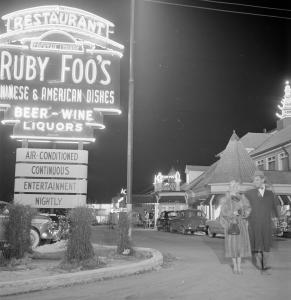Photographie de soir de l’extérieur du restaurant. Les enseignes au néon à gauche et au fond de l’image sont illuminées. À droite, un couple sort du stationnement.