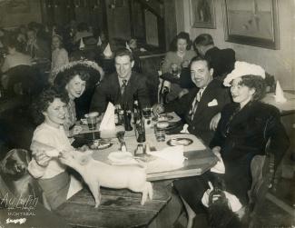 Trois femmes et deux hommes sont attablés dans un restaurant. La femme de gauche donne le biberon à un porcelet à l’avant-plan. À l’arrière-plan, on voit d’autres clients attablés.