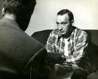 Photographie en noir et blanc, un homme à gauche est dos à l’objectif alors que celui de droite est de face. Il porte une chemise à carreaux.