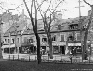 Photographie d'une enfilade de bâtiments en pierre abritant des commerces, dont « Laundry » et « A. Martin Bijoutier & Opticien ».