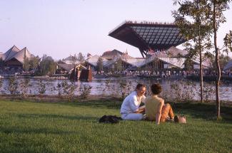 Photo couleur montrant le pavillon du Canada en arrière-plan. En avant-plan, un jeune couple est assis dans l'herbe.