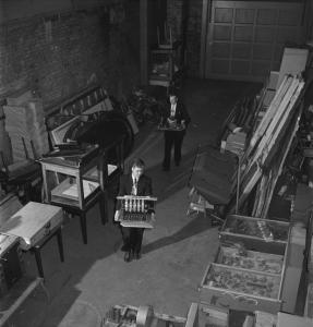 Intérieur d’un entrepôt. Deux hommes tiennent des machines dans leurs mains. De chaque côté, du matériel est empilé.