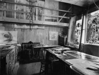 Grande pièce d'un atelier. Plusieurs œuvres sont accrochées au mur ou déposées sur des tables.