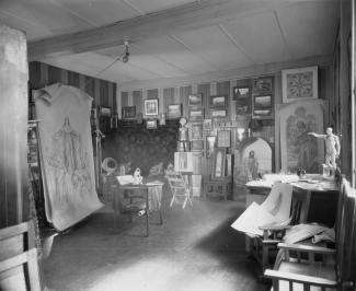 Table de travail au premier plan, avec matériel artistique. Au deuxième plan, des vitraux et des esquisses sont accrochées au mur.