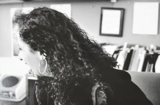 Une femme, au longs cheveux noirs, de profil. En arrière-plan, des cadres accrochés au mur et des boites de dossiers.