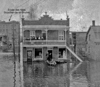 Photo en noir et blanc montrant une rue inondée avec une maison à deux étages en plein centre. Trois hommes sont dans une chaloupe et quelques personnes sont sur le balcon du haut.