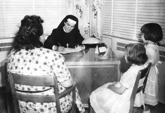 Marie Gérin-Lajoie à un bureau, en entrevue de service social avec une femme et deux jeunes filles.